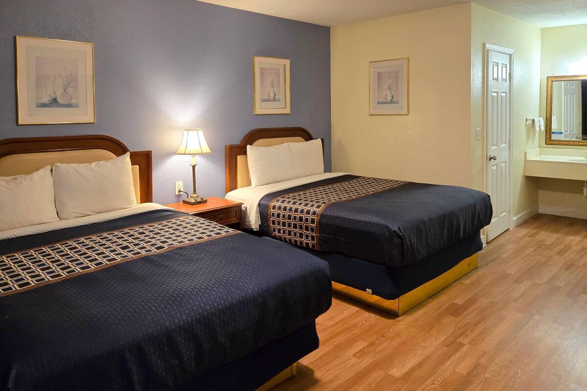 A hotel room with two beds, a nightstand, a lamp, and a bathroom with a mirror and sink.