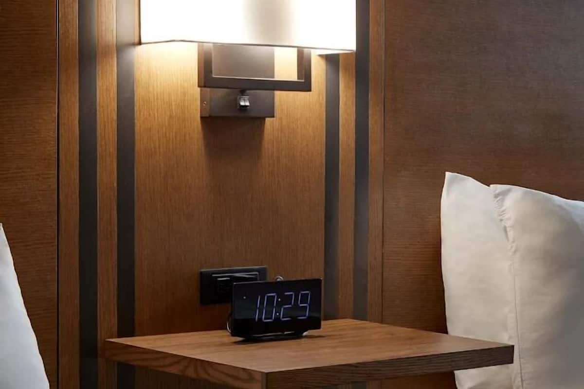 A hotel room with a bedside table, a digital clock, a wall-mounted lamp, and a bed with pillows.