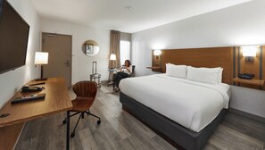 A hotel room with a bed, desk, chair, and a person sitting at the desk.