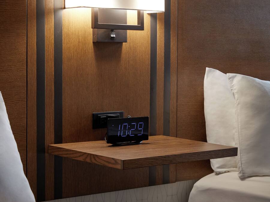 Una habitación de hotel con una mesita de noche, un reloj digital y una lámpara fijada a la pared.