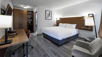 A hotel room with a bed, desk, chair, and a small table with a lamp.