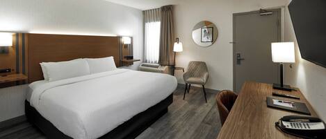 A hotel room with a large bed, a desk with a telephone, a chair, a desk lamp, a clock, and a door.
