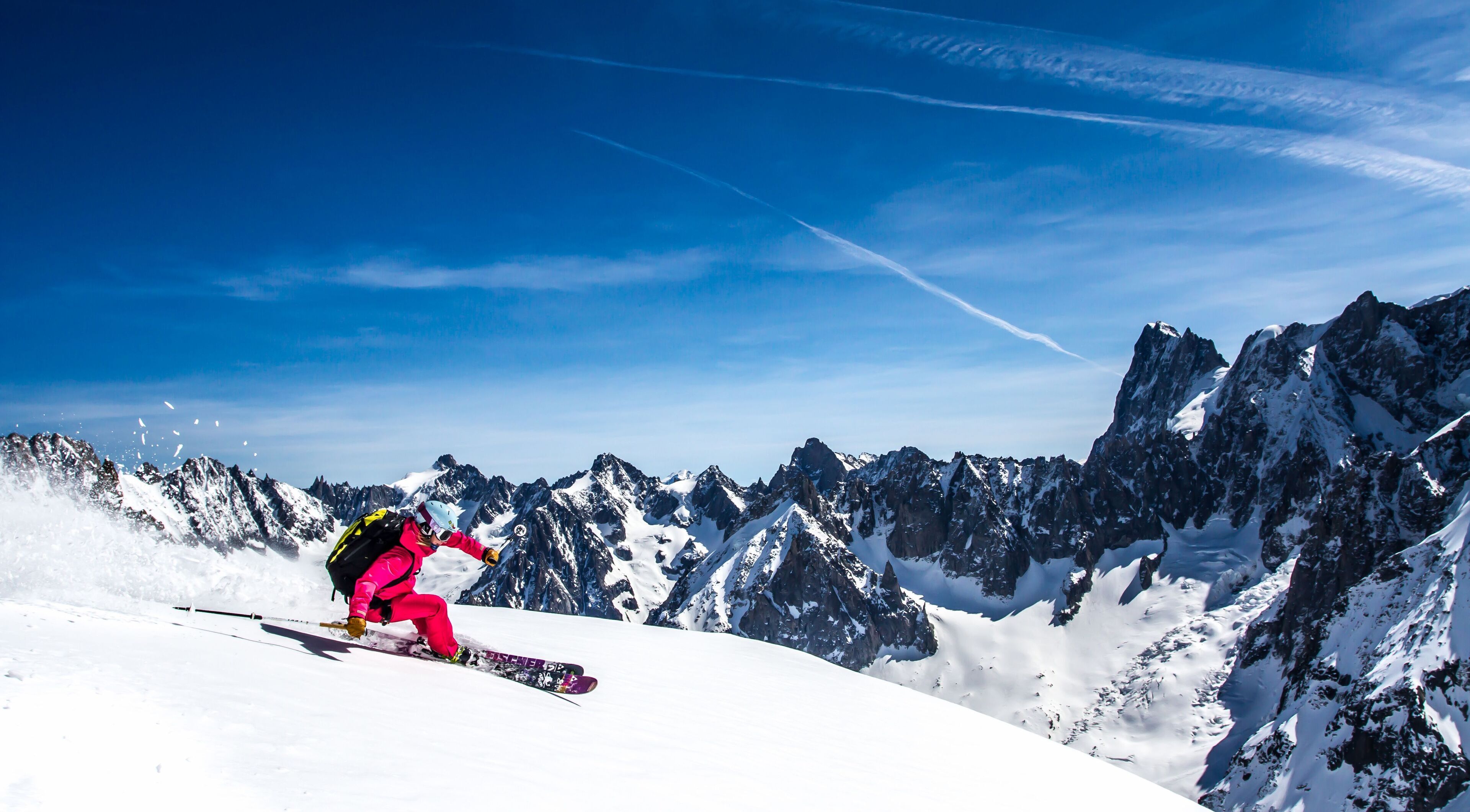 A snowboarder in pink gear on a snowy mountain slope with a clear blue sky and rugged mountain peaks in the background.