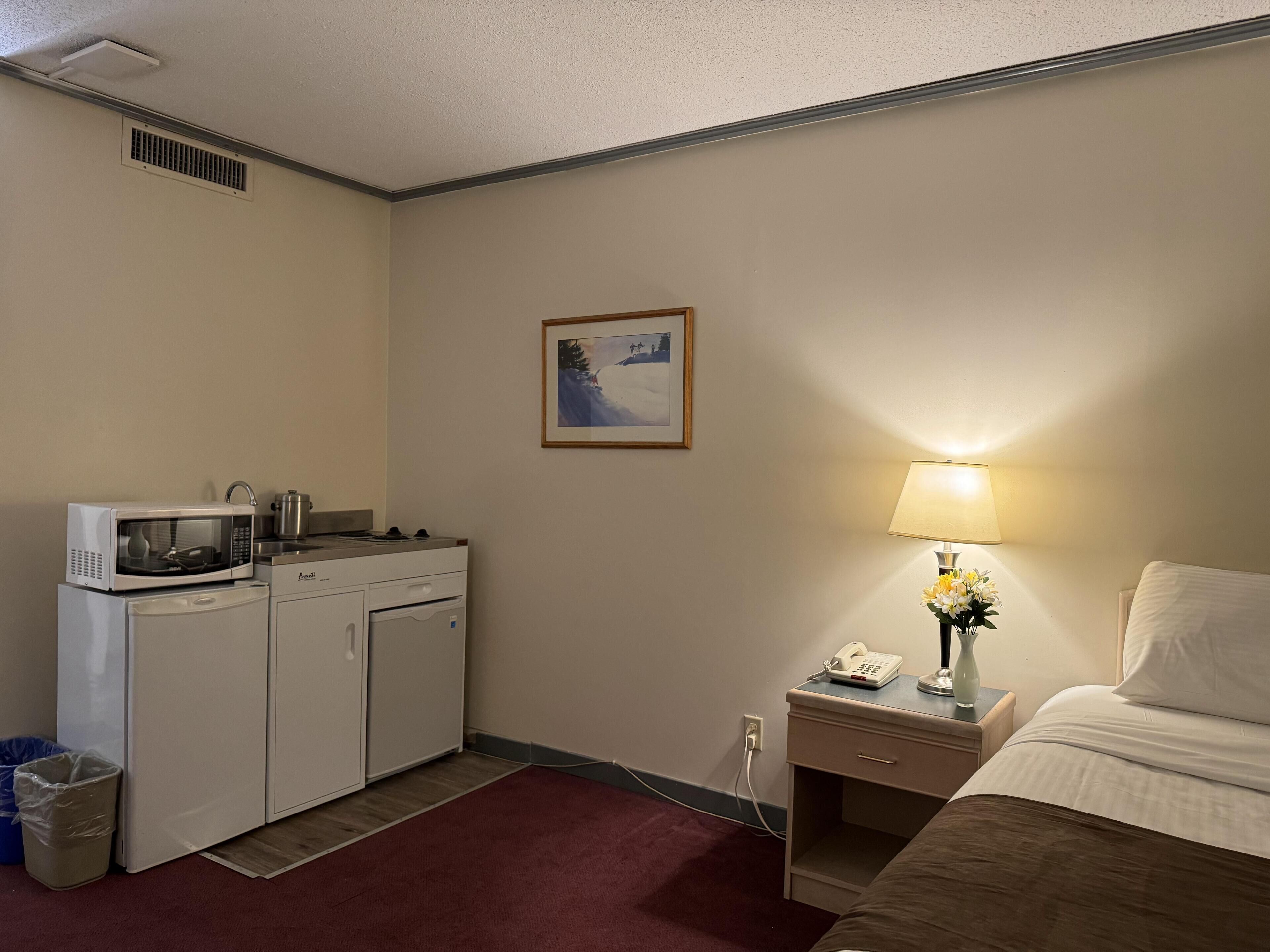 A hotel room with a kitchenette, a bed, a nightstand with a lamp, a picture on the wall, and a trash bin.