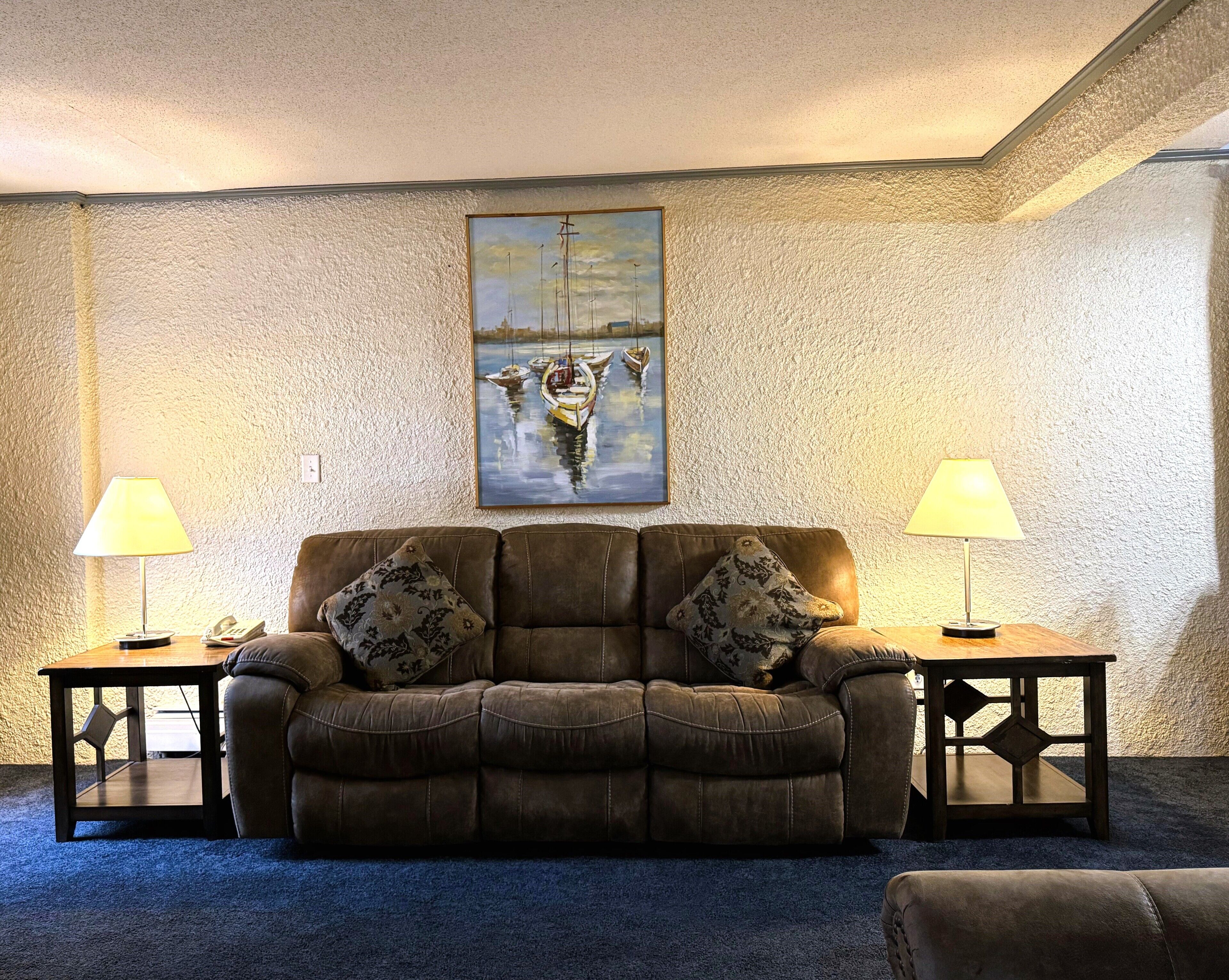 A cozy living room with a brown leather sofa, two matching lamps, and a painting of boats on the wall.