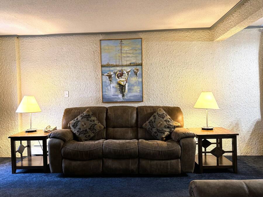 A cozy living room with a brown leather sofa, two matching lamps, and a painting of boats on the wall.
