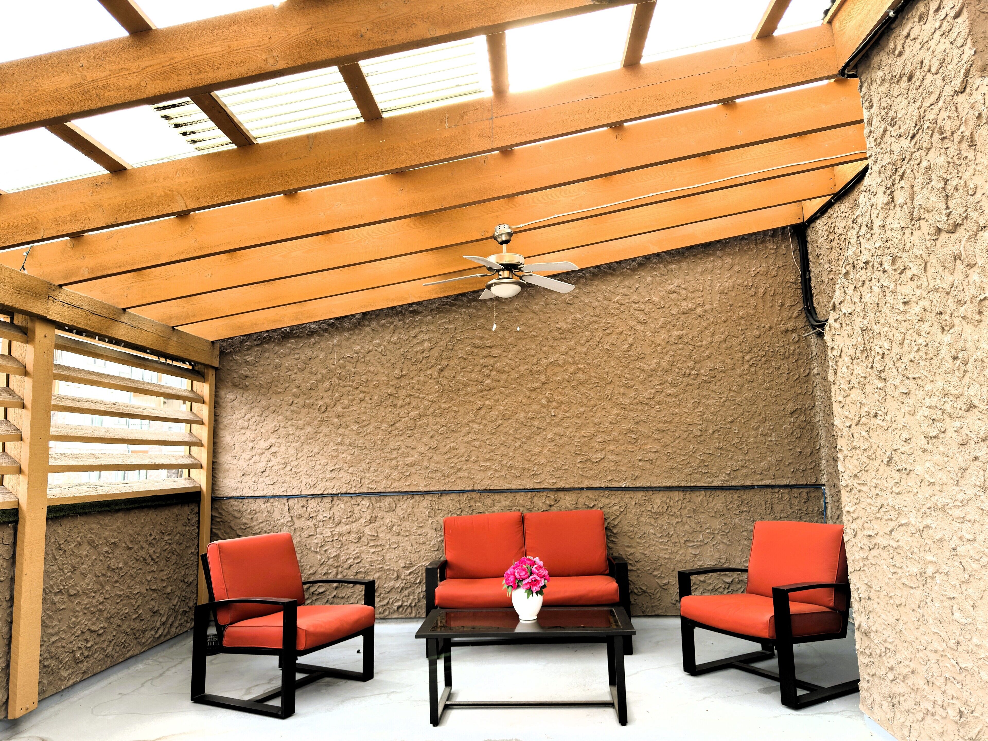 A covered patio area with a wooden ceiling, a ceiling fan, and three red chairs around a small table with a vase of flowers.