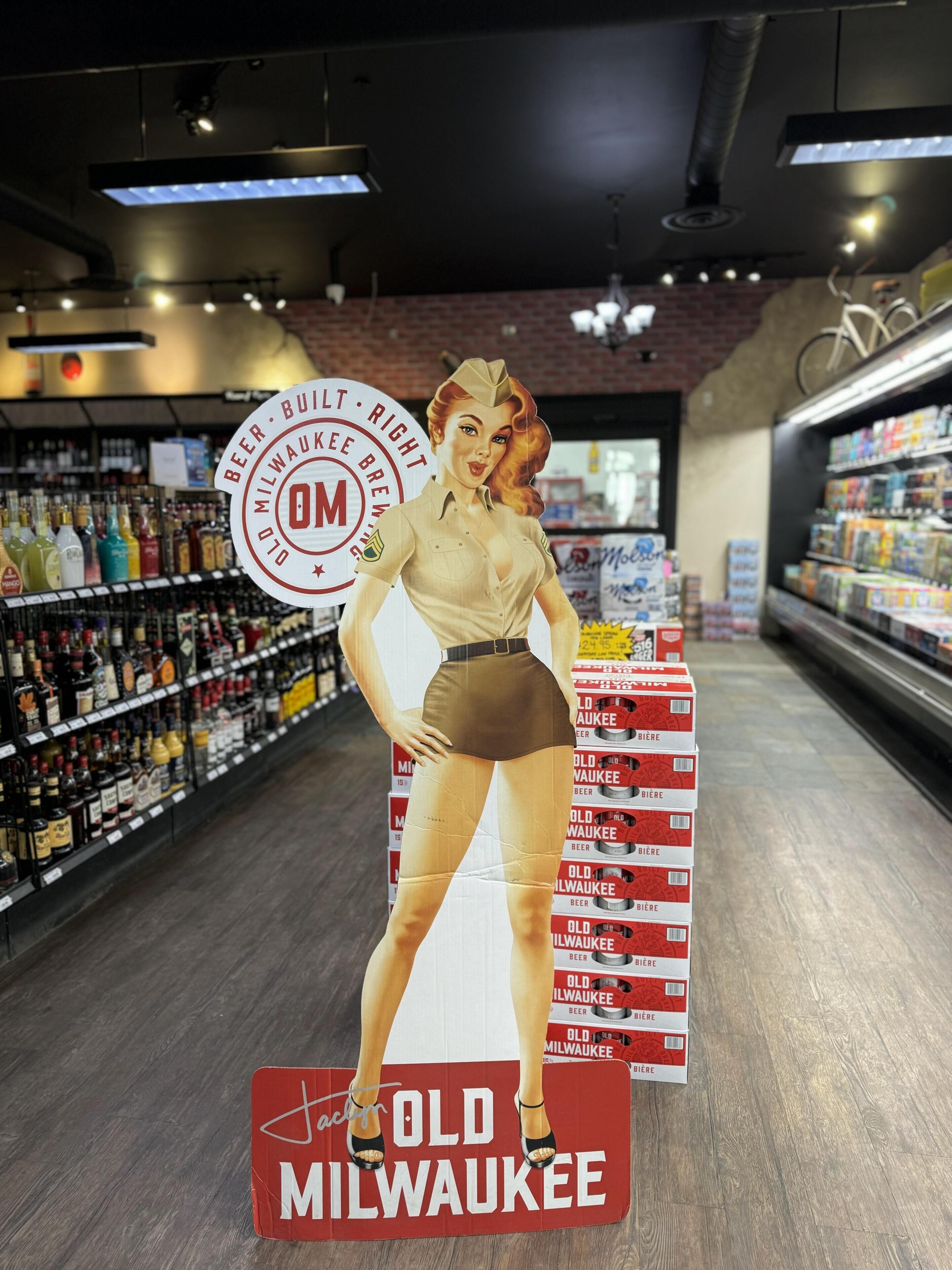 Een etalagepop in een gangpad tussen schappen vol drankflessen en stapels bierkartons met de tekst 'Old Milwaukee'.
