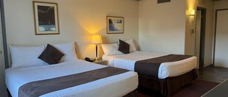 A hotel room with two beds, each with white linens and brown headboards. There are two bedside tables with lamps, and the walls are adorned with framed pictures.
