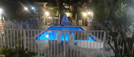 Una piscina con cerco blanco, rodeada de plantas en macetas y mobiliario exterior.