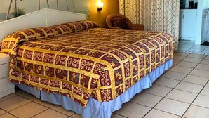 A hotel room with a large bed, a nightstand, a wall-mounted lamp, a chair, and a view of palm trees.