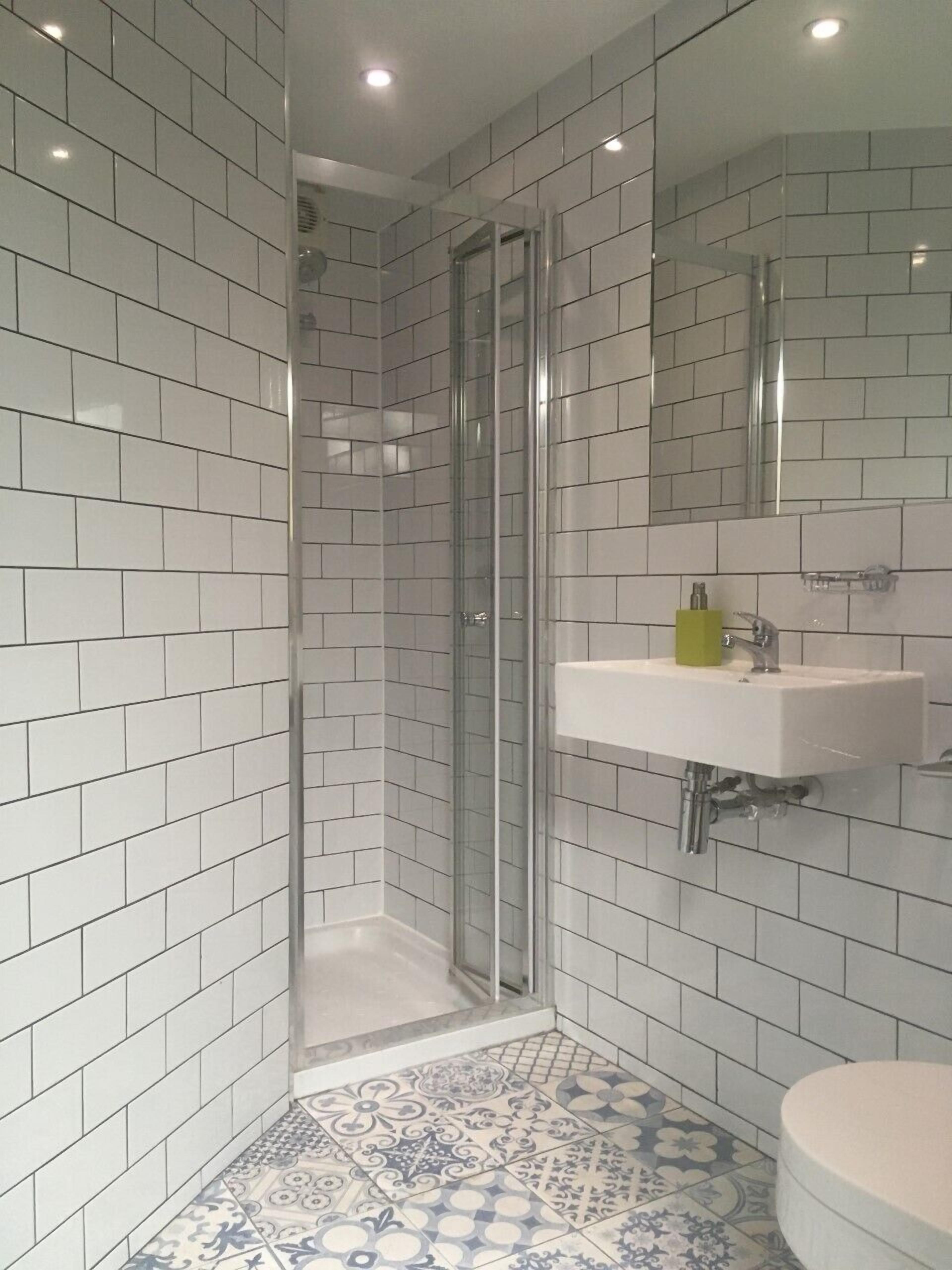 A modern bathroom with white subway tiles, a glass shower enclosure, a white sink with a green soap dispenser, and patterned floor tiles.