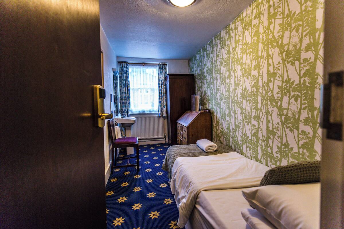 A hotel room with a bed, a chair, a small table, a radiator, and a window with curtains.