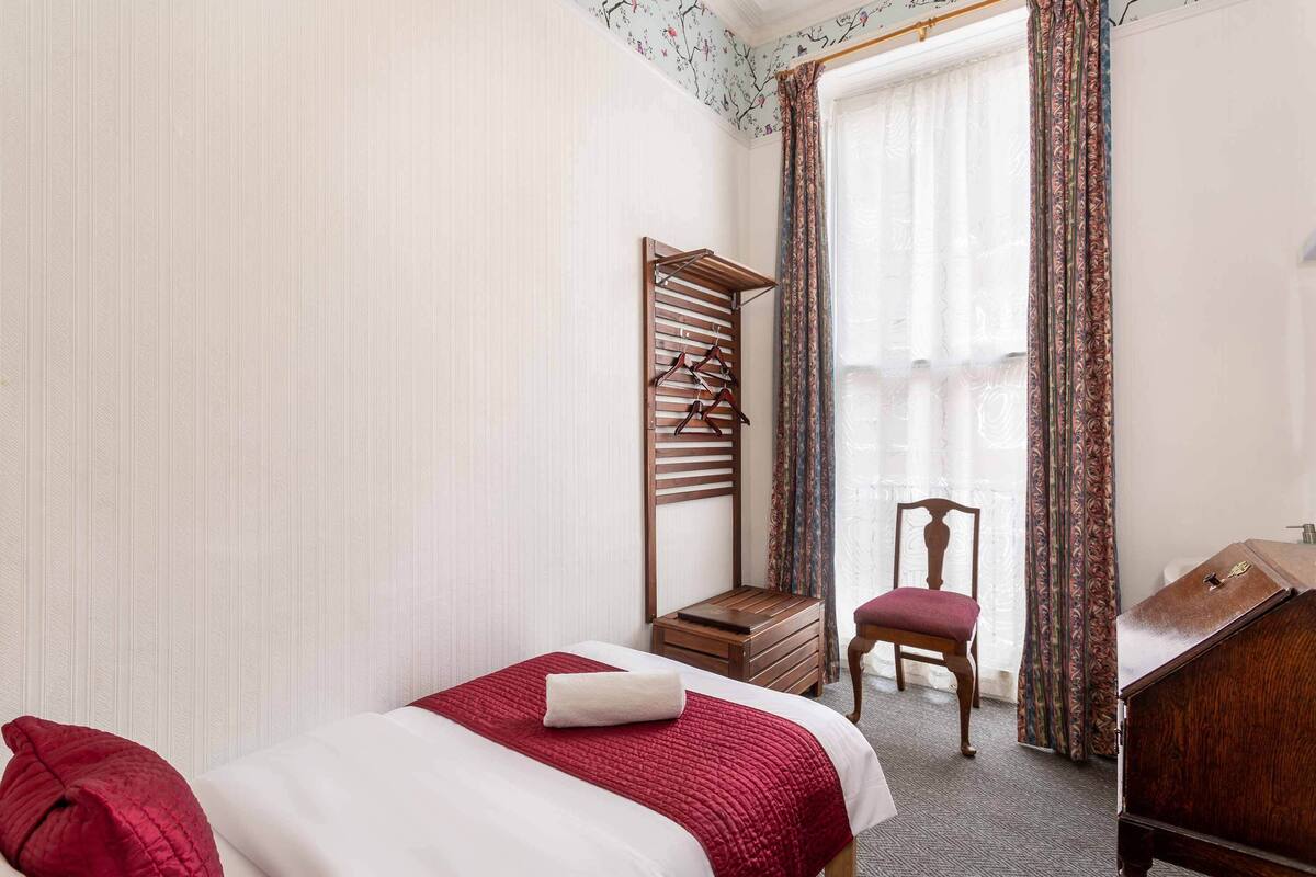 A hotel room with a bed, a chair, a wooden side table, a mirror, and a coat rack.