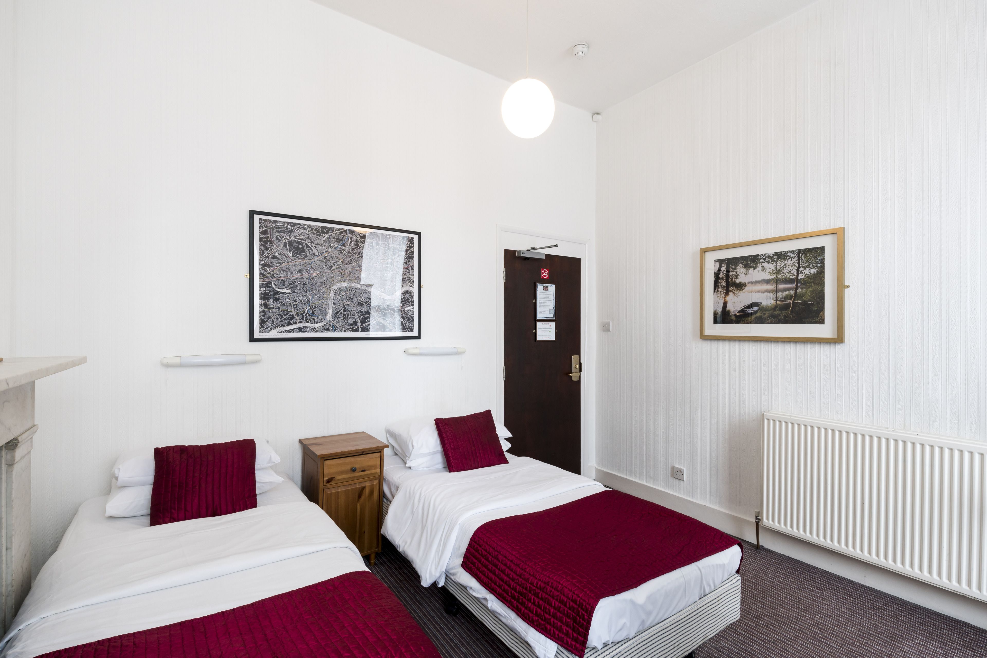 A hotel room with two beds, a wooden nightstand, a white radiator, and two framed pictures on the walls.