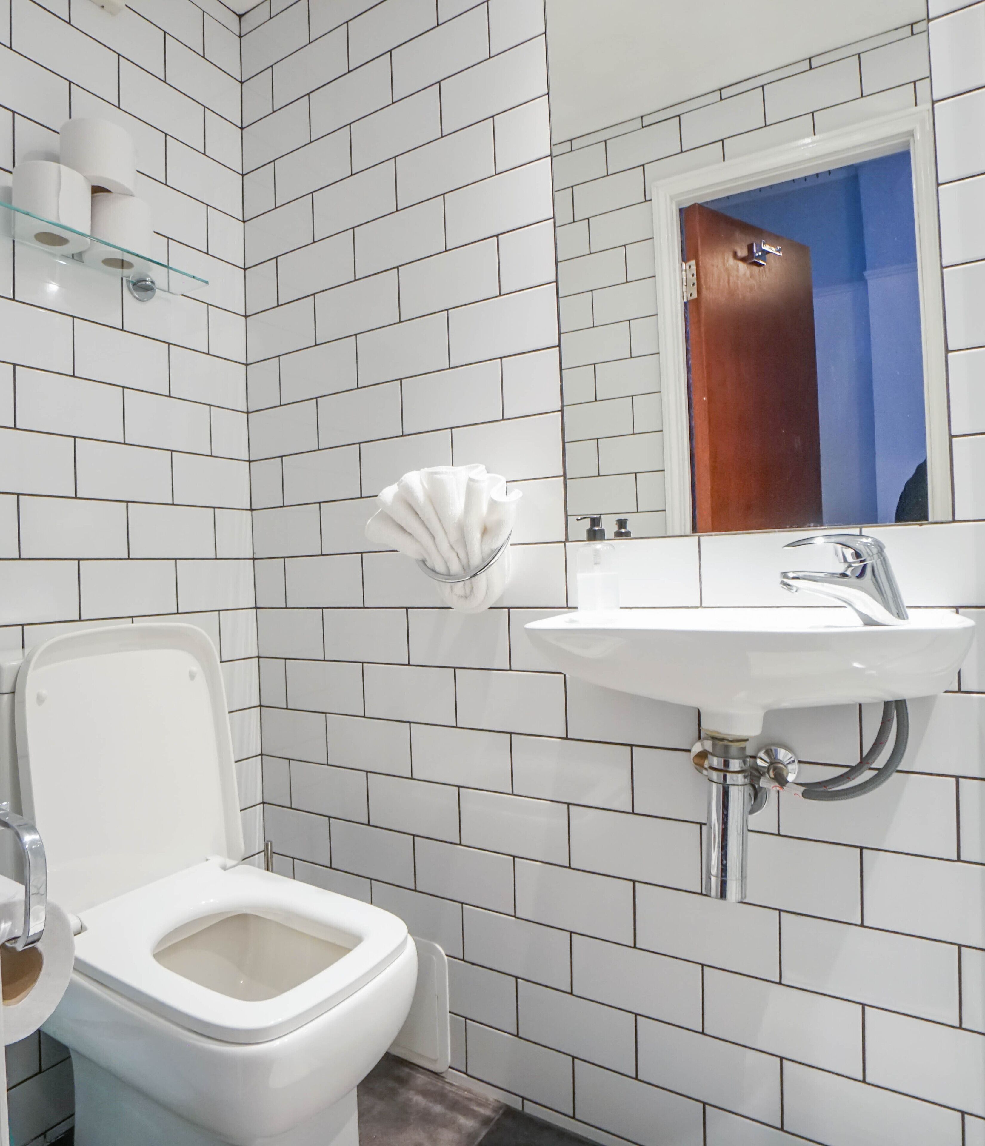 A bathroom with white tiled walls, a white toilet, a white sink with a mirror above it, and a glass shelf with toiletries.