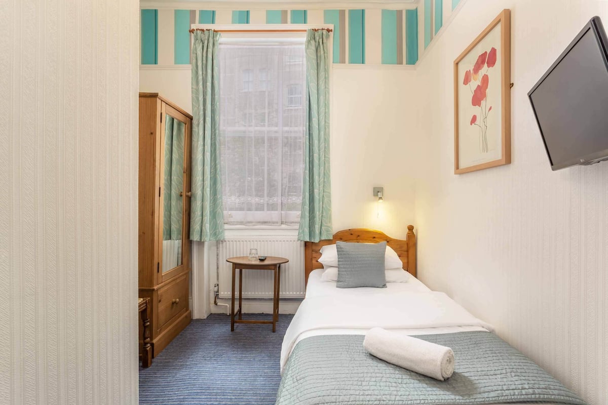 A small, neatly arranged bedroom with a single bed, a wooden wardrobe, a small table, and a window with curtains.