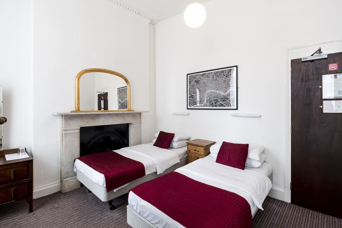 A hotel room with two single beds, a fireplace, a mirror, a framed abstract painting, a wooden nightstand, and a door with a sign.