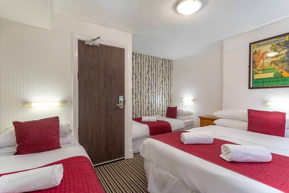 A hotel room with two beds, each with red bedding and white pillows. There is a wooden door, a patterned curtain, and a poster on the wall.