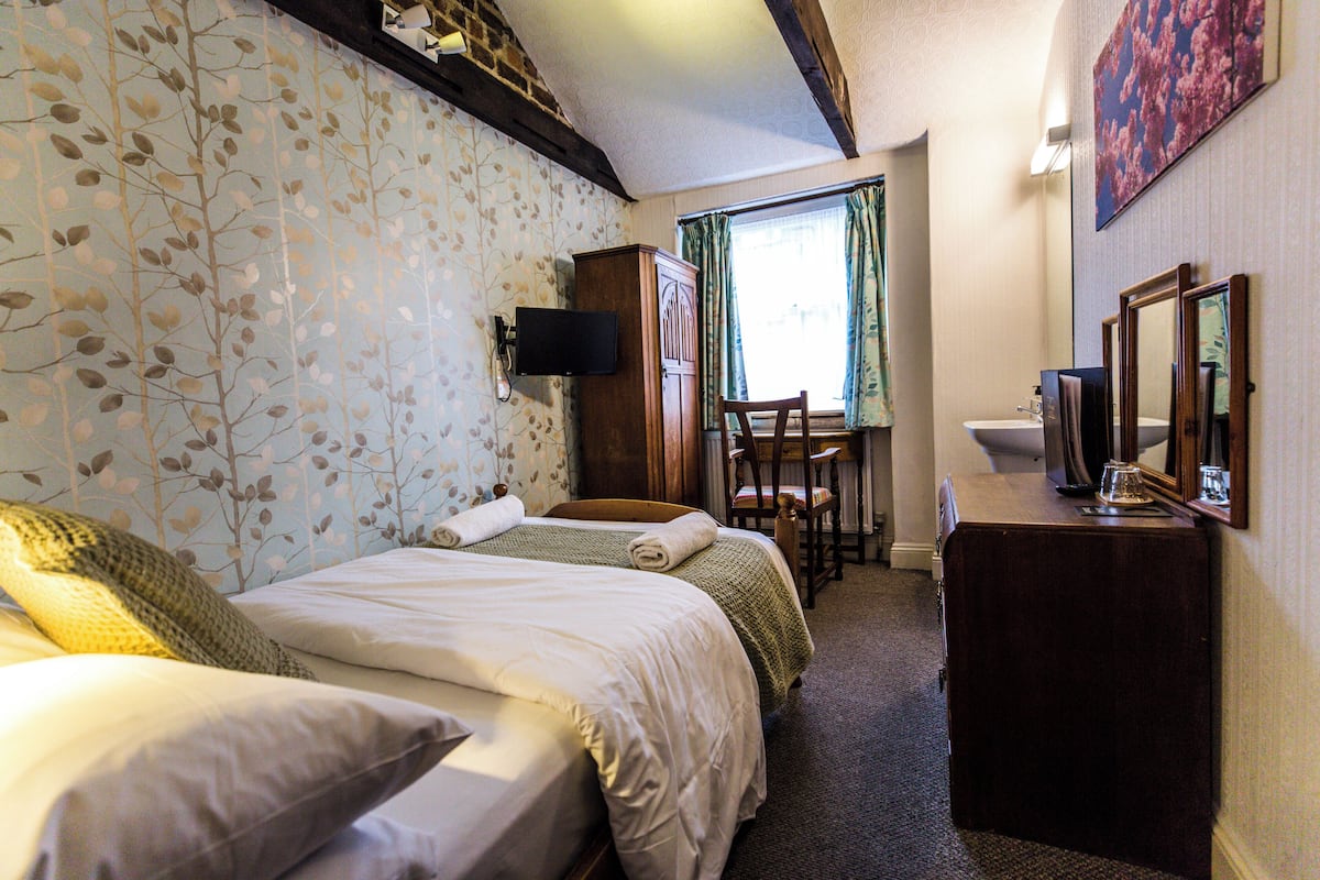 A hotel room with two beds, a desk, a chair, a wardrobe, and a bathroom with a mirror and sink.