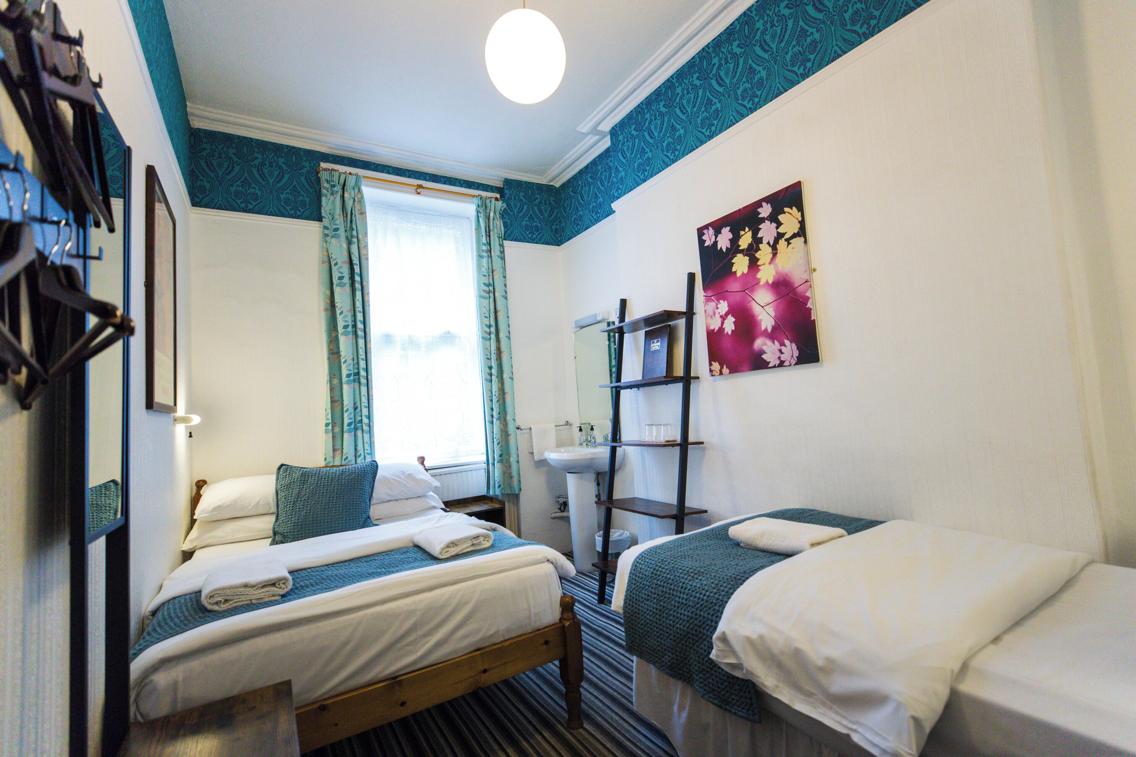 A hotel room with two single beds, a window with curtains, a sink, and a shelf with toiletries.