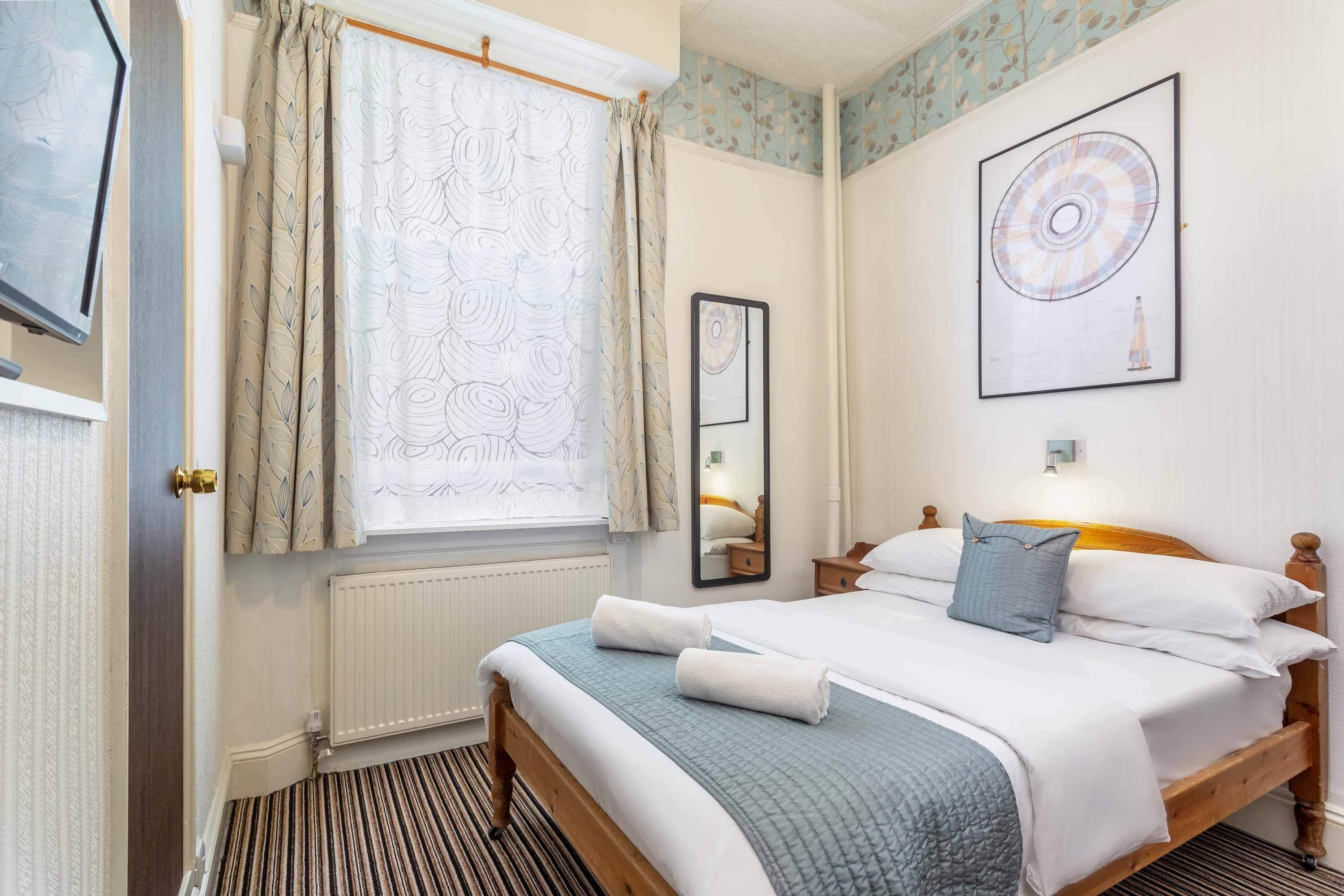 Een net opgemaakt bed met een blauwe deken en witte kussens, een houten hoofdbord, een raam met gordijnen met een patroon, een spiegel aan de muur en een kunstwerk in een lijst boven het bed.