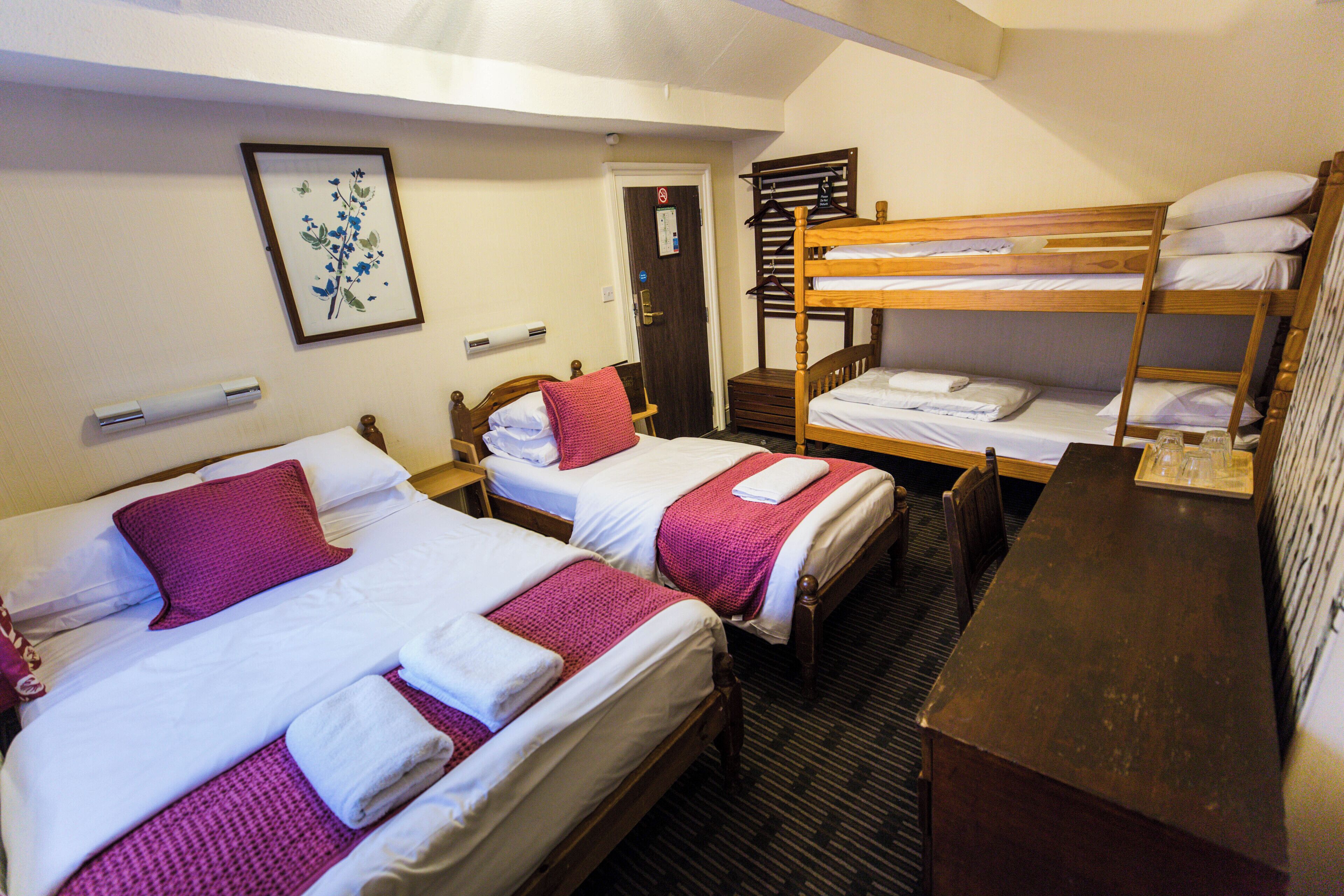 A hotel room with two single beds, a bunk bed, a wooden desk, and a framed floral picture on the wall.