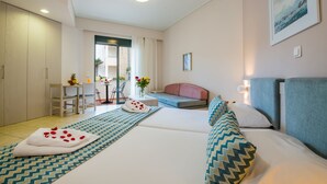 A hotel room with a bed, a sofa, a desk with a chair, a small table with a vase of flowers, and a view of the outdoors through a glass door. - Creta Palm Hotel (Chania)