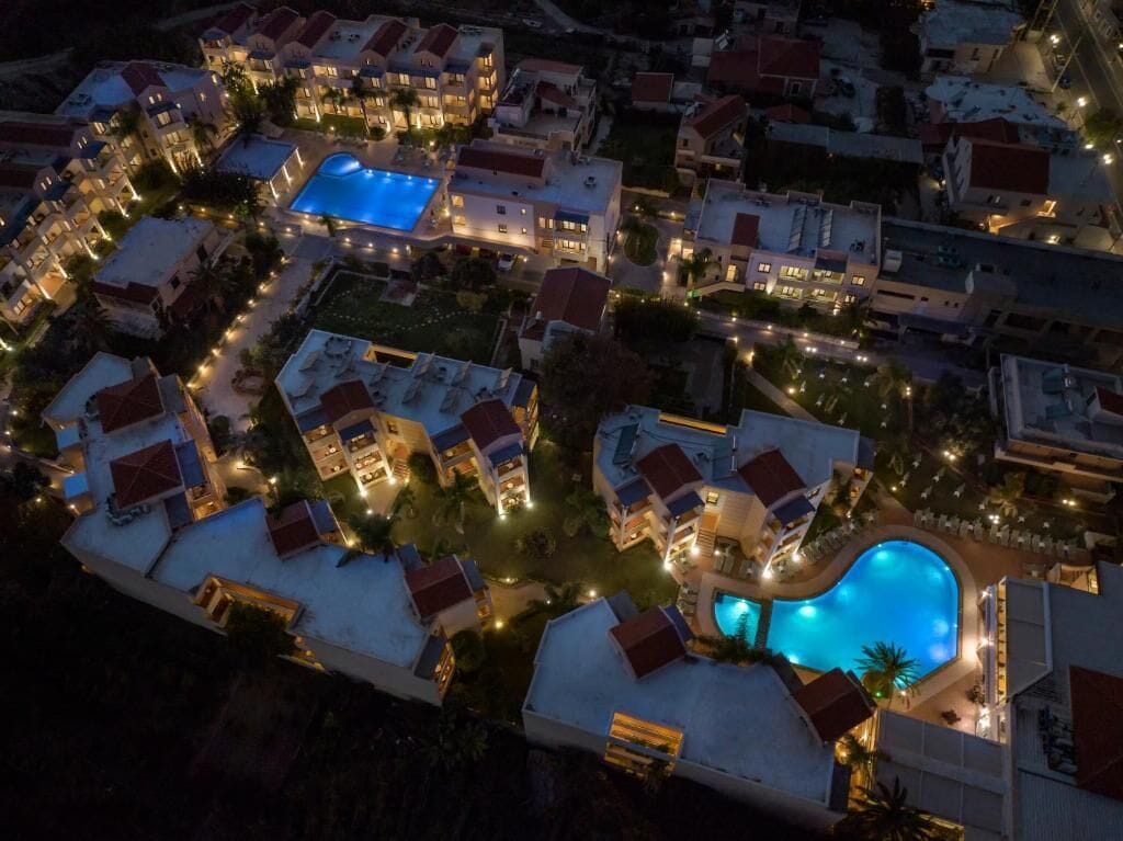 An aerial view of a resort with multiple buildings, a large swimming pool, and well-lit pathways.