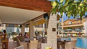 An outdoor seating area with wicker furniture and a pool in the background. - Creta Palm Hotel (Chania)