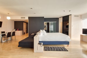 A modern hotel room with a bed, desk, and chair, a small kitchenette, and a TV.