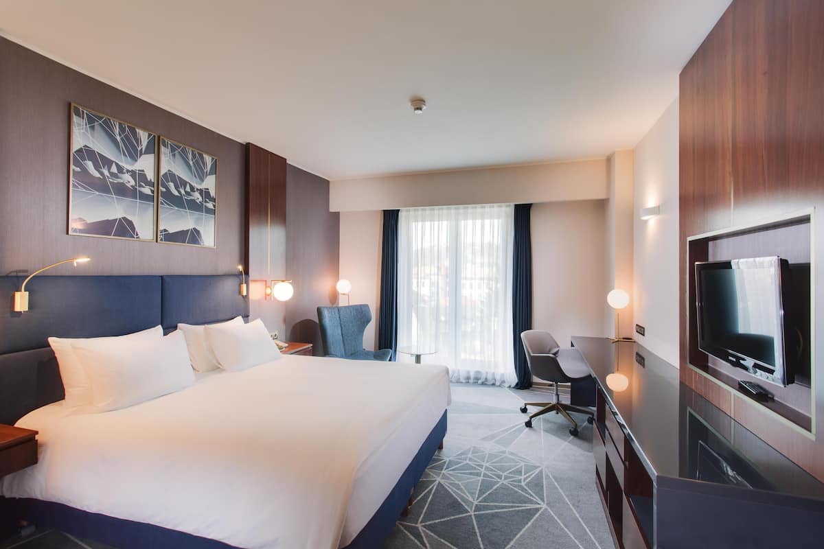 Une chambre d’hôtel avec un grand lit, un bureau avec une télévision, une chaise et une fenêtre avec des rideaux.