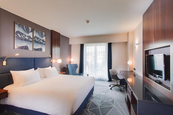 Habitación de hotel con una cama grande, un escritorio con televisor, una silla y un ventanal con cortinas.