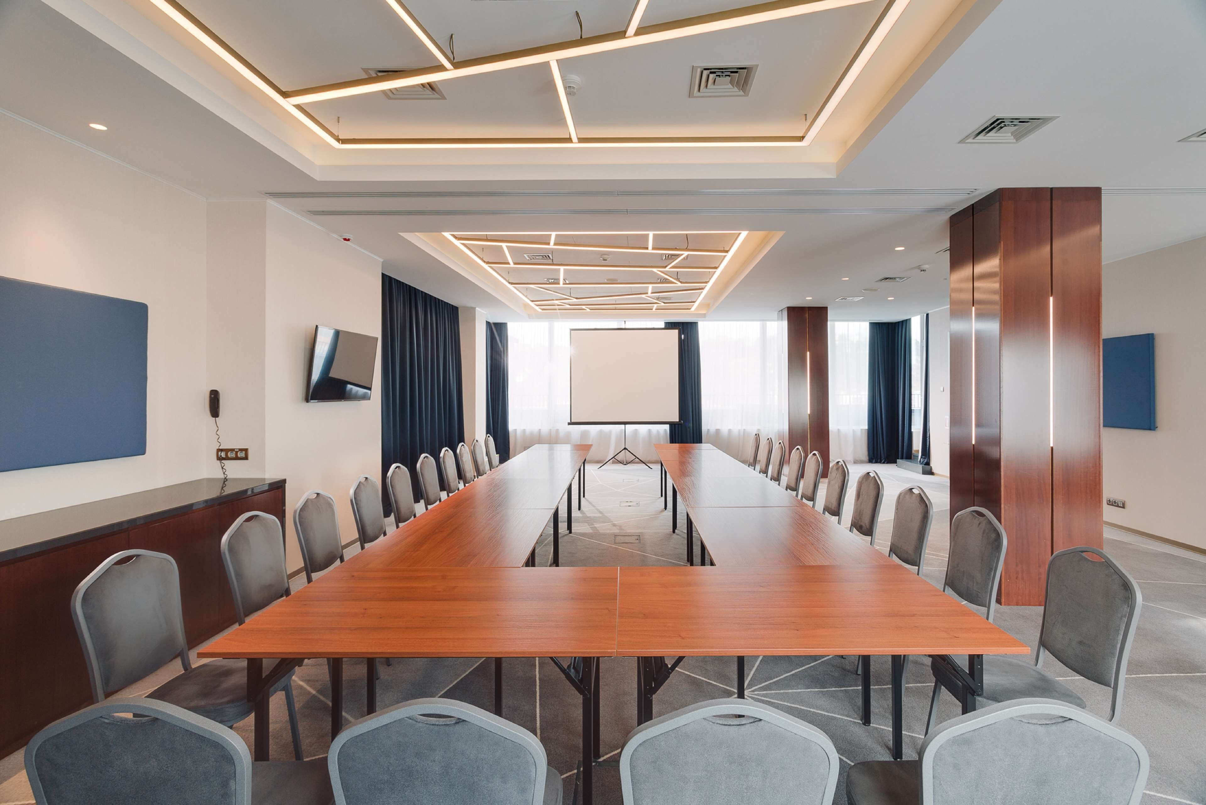 Un salón de conferencias con una larga mesa de madera, sillas tapizadas en gris, una pizarra y una pantalla para proyector.