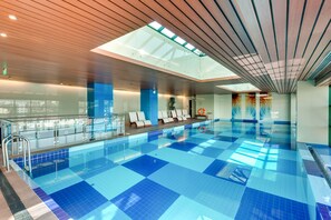 Piscina interna com azulejos azuis translúcidos, teto de vidro e design moderno.