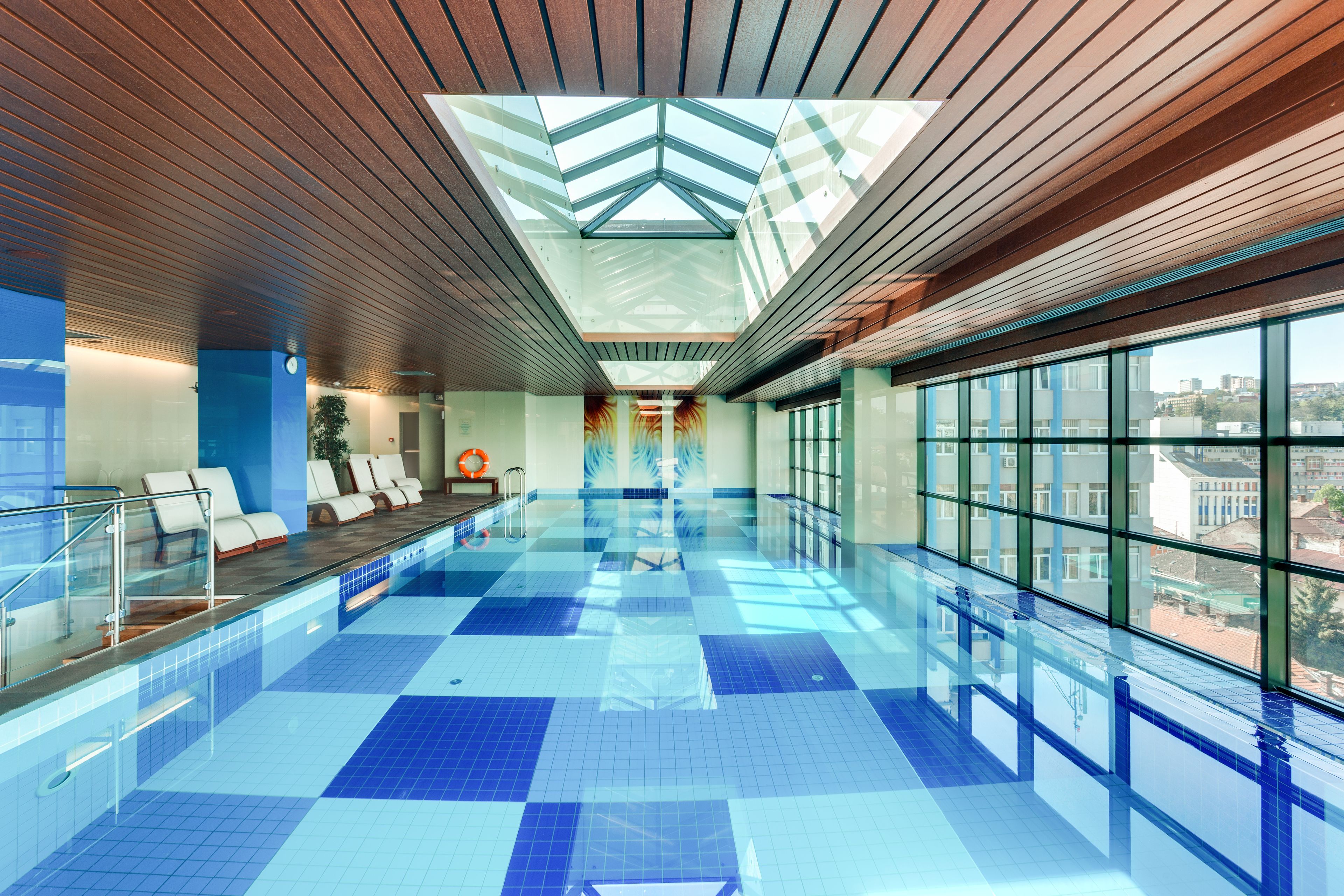A modern indoor swimming pool with a skylight ceiling, clear glass walls, and a view of the city.