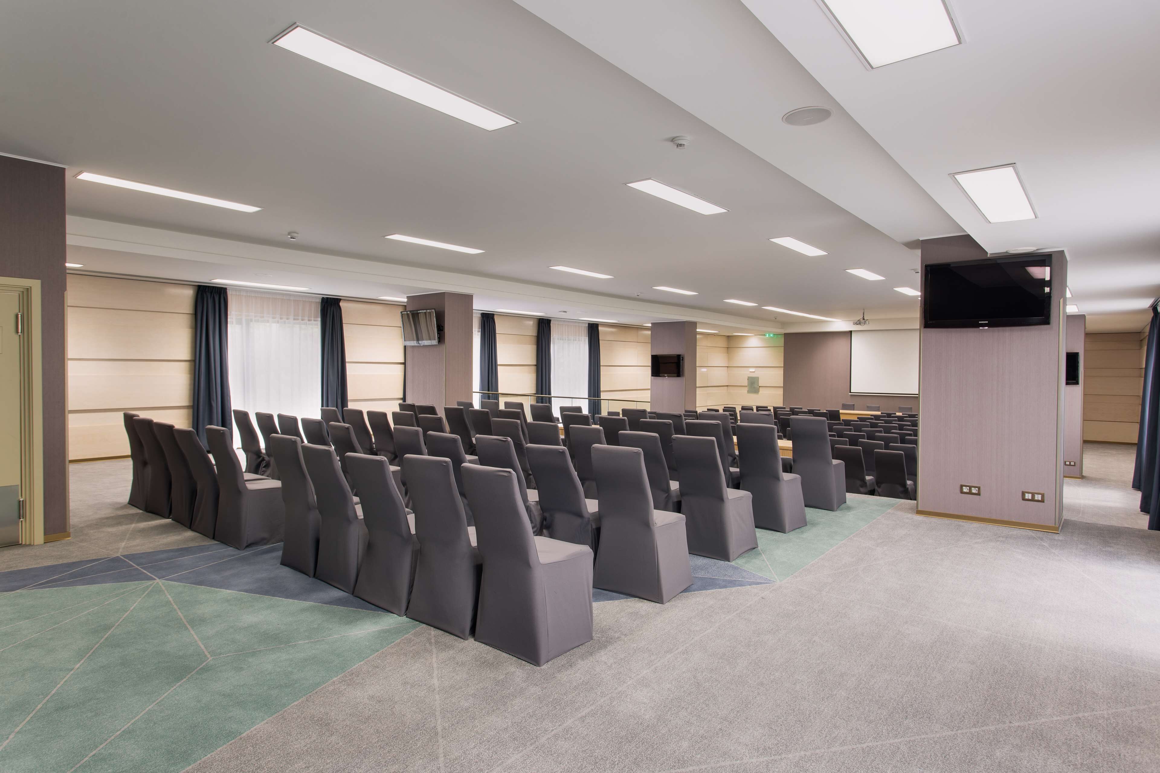 A conference room with rows of chairs, a large screen, and a flat-screen TV mounted on a wall.