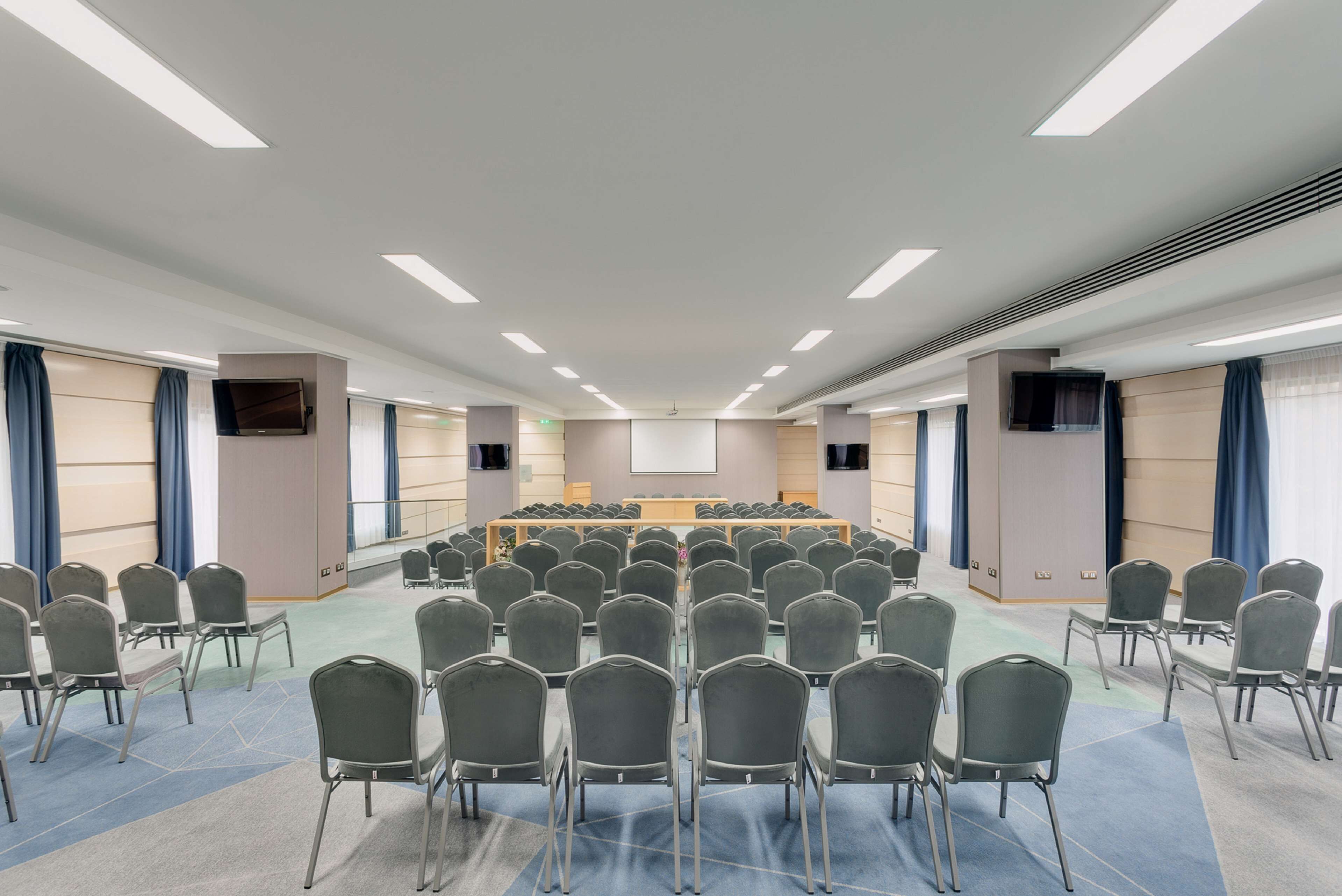 Un salón de conferencias con filas de sillas orientadas hacia un escenario central, equipado con un proyector y una pantalla.
