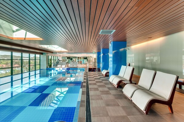 A modern indoor swimming pool with clear blue tiles, wooden ceiling slats, and lounge chairs along the side.