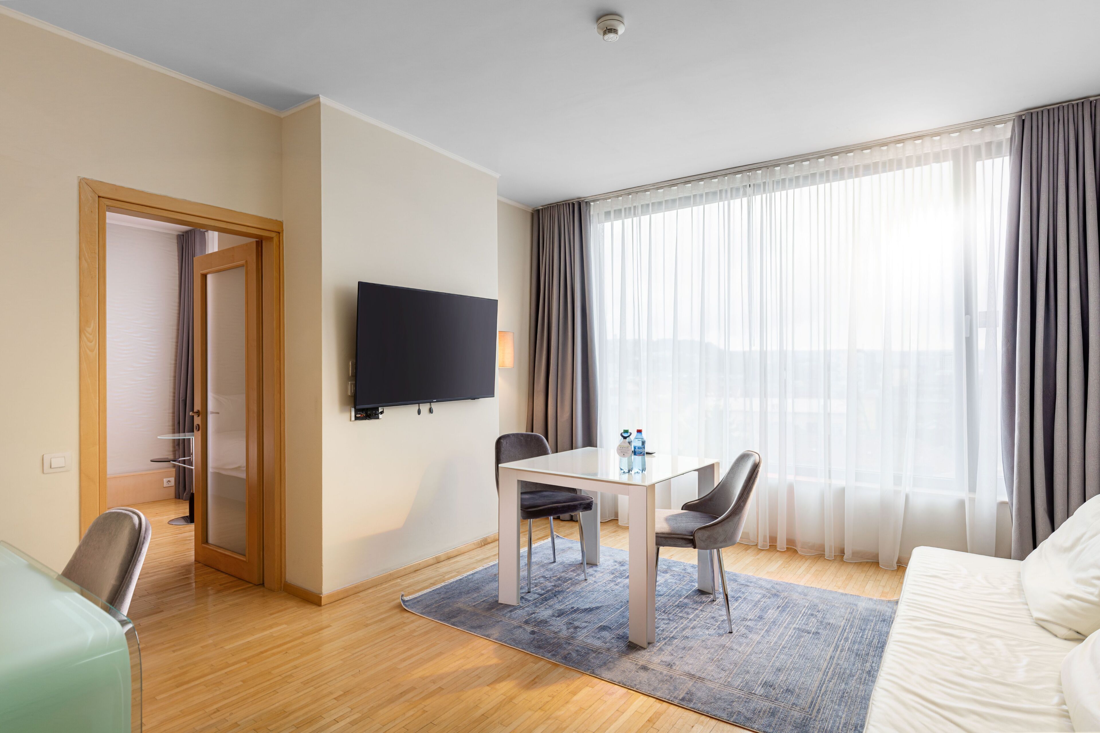 Une chambre d’hôtel avec un plancher en bois, une table à manger avec des chaises, un téléviseur fixé au mur et une grande fenêtre avec des rideaux.