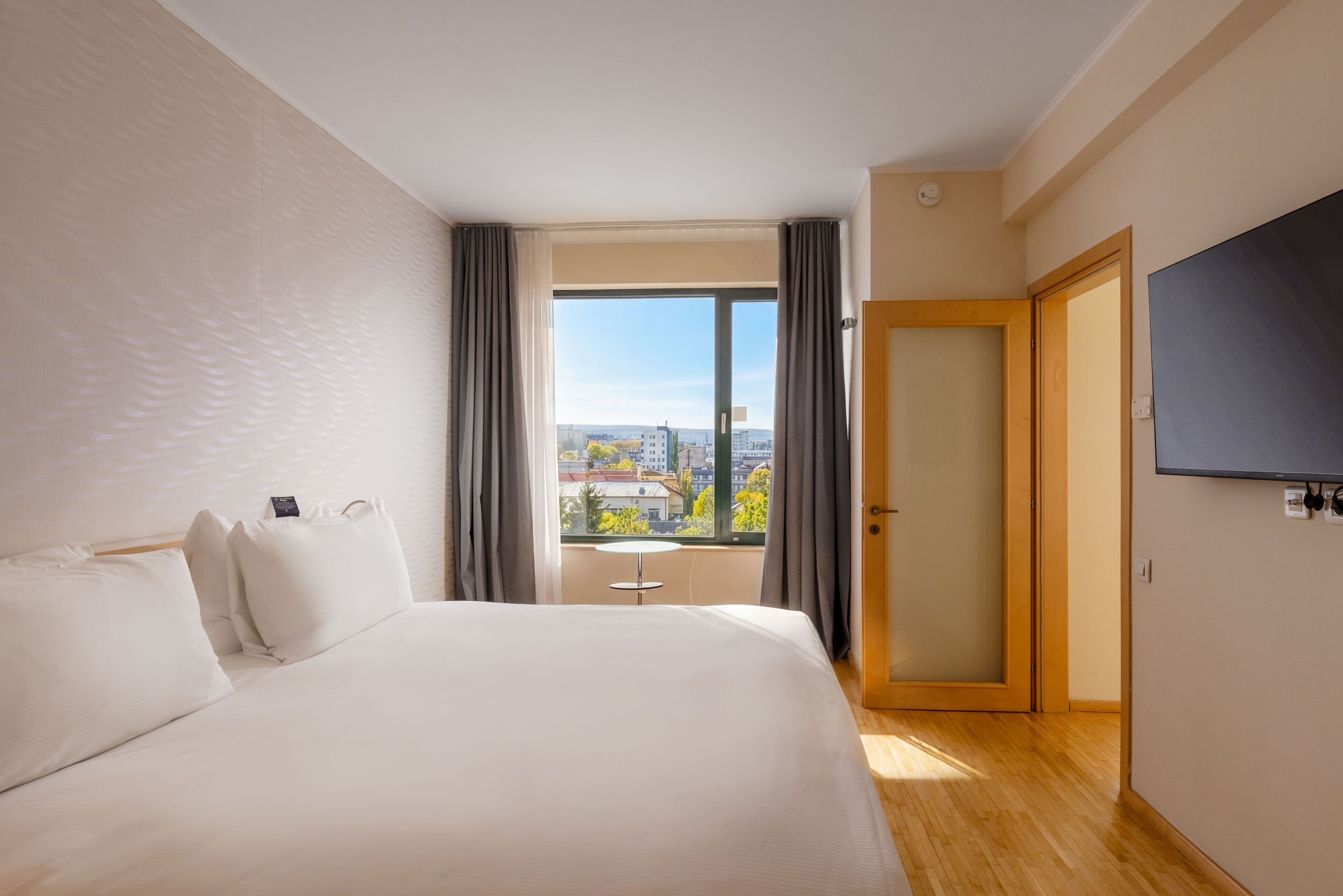 A hotel room with a large bed, a TV mounted on the wall, a window with curtains, and a view of the cityscape.