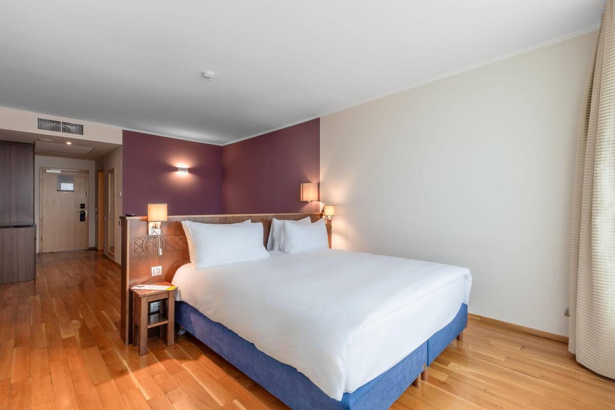 Une chambre d’hôtel avec un grand lit, deux lampes de chevet, une petite table et une porte donnant sur un couloir.