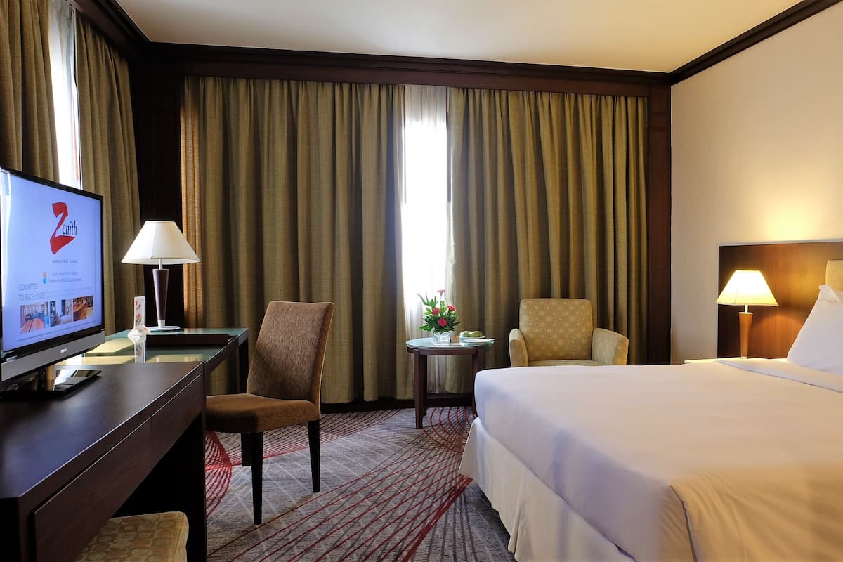 A hotel room with a bed, two chairs, a desk with a computer, a lamp, and a TV displaying the Zenith logo.