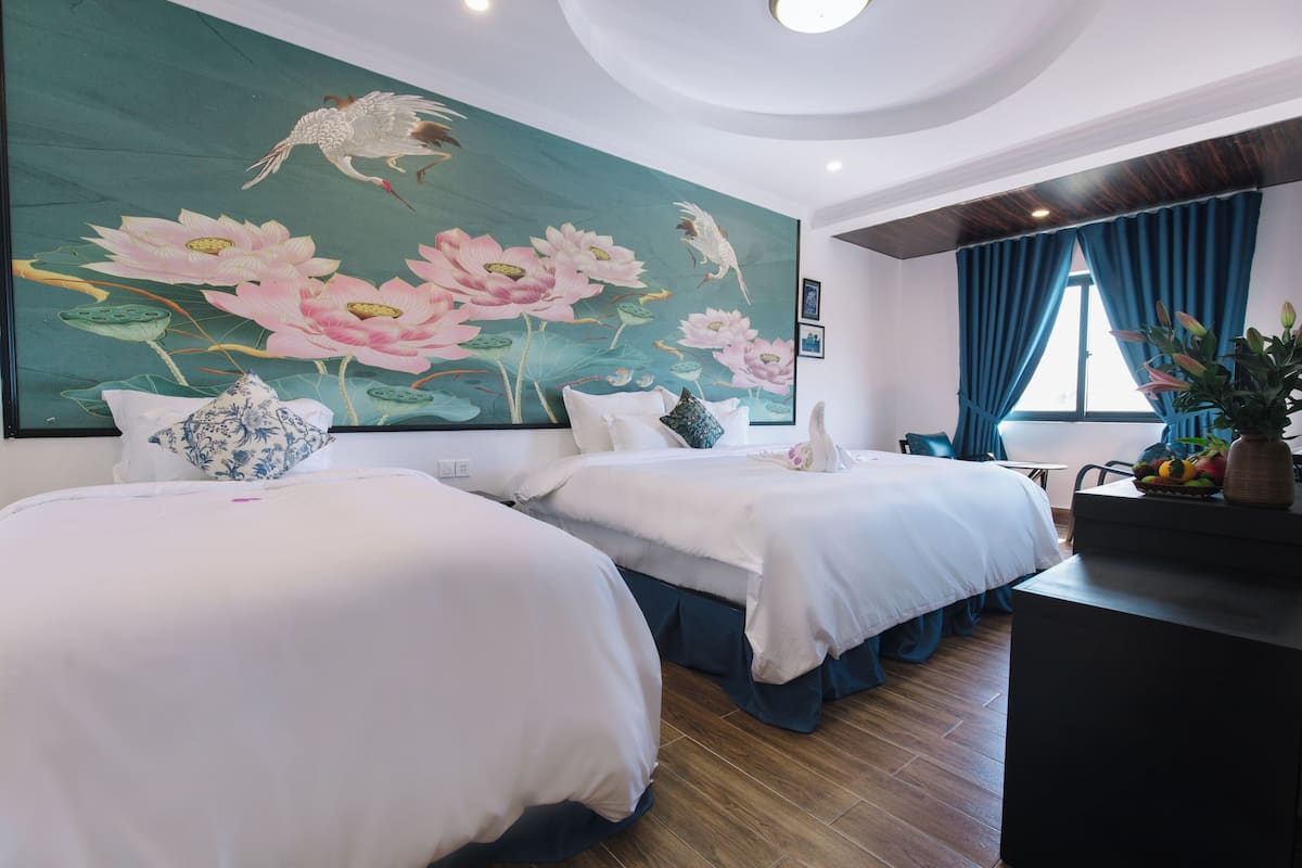 A hotel room with two beds, a large floral mural, a desk with a vase, and a window with curtains.