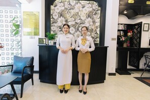 A reception area with a floral backdrop, a reception desk, a seating area with a blue chair, and a bookshelf with books and decorative items. - Orchid Hotel (Hue)
