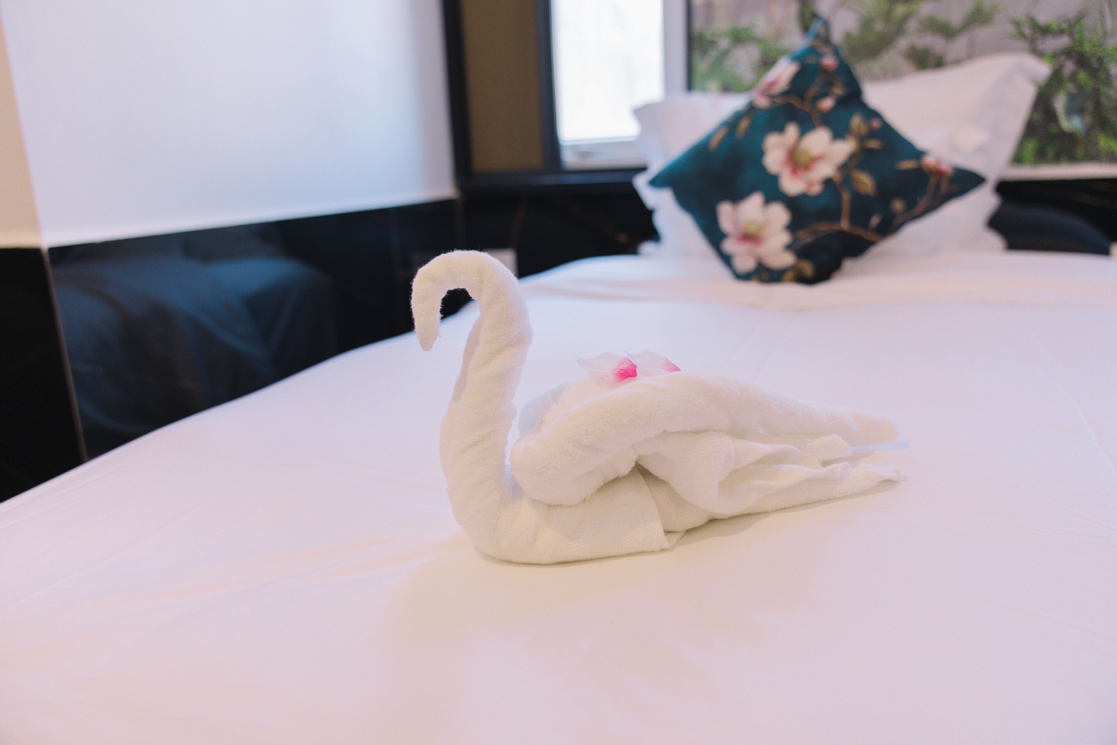 Una toalla con forma de cisne sobre una cama con un cojín estampado y una ventana al fondo.