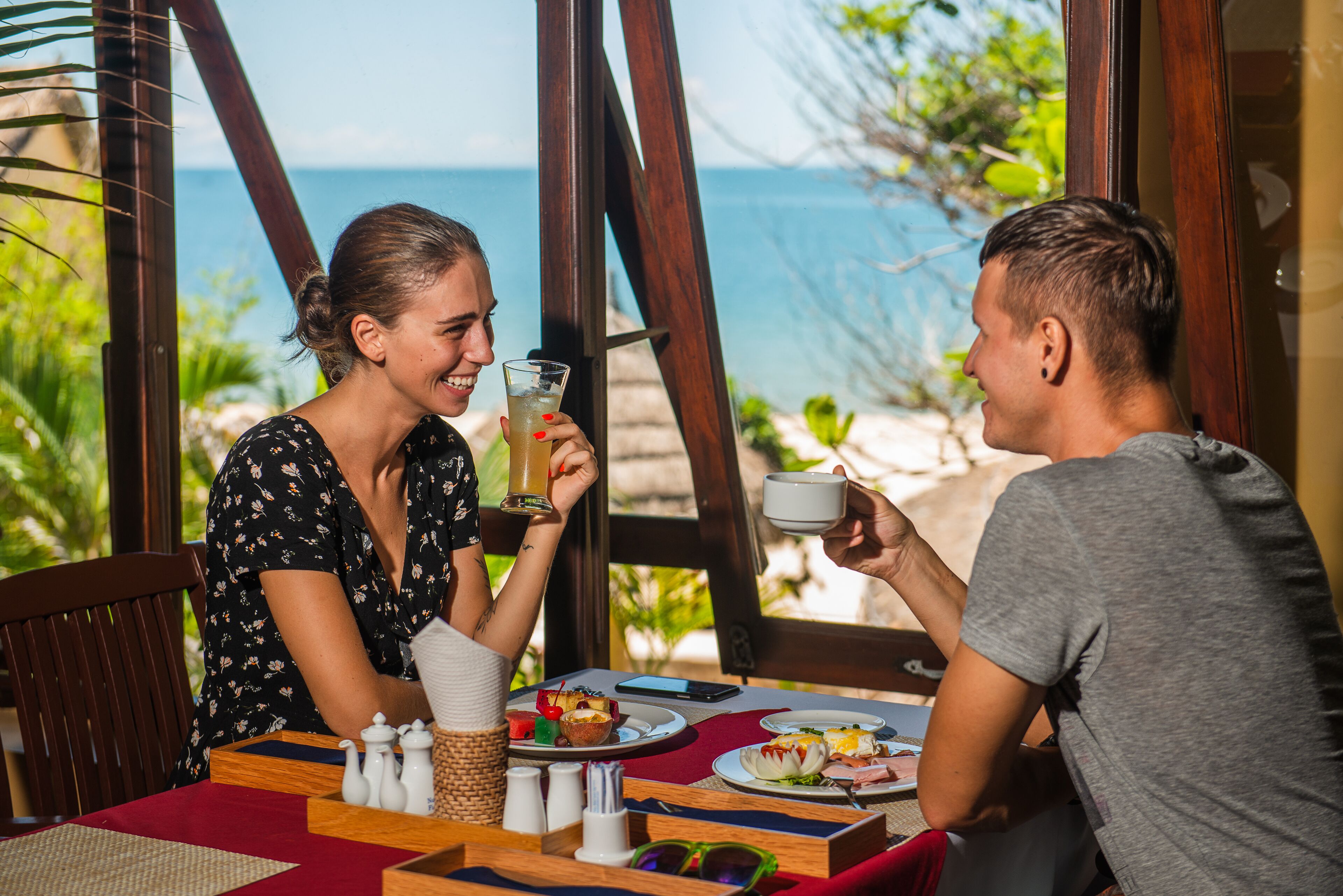 Una mesa de comedor con platos de comida, un vaso de bebida y una taza, frente a una estructura de madera y vistas al mar.