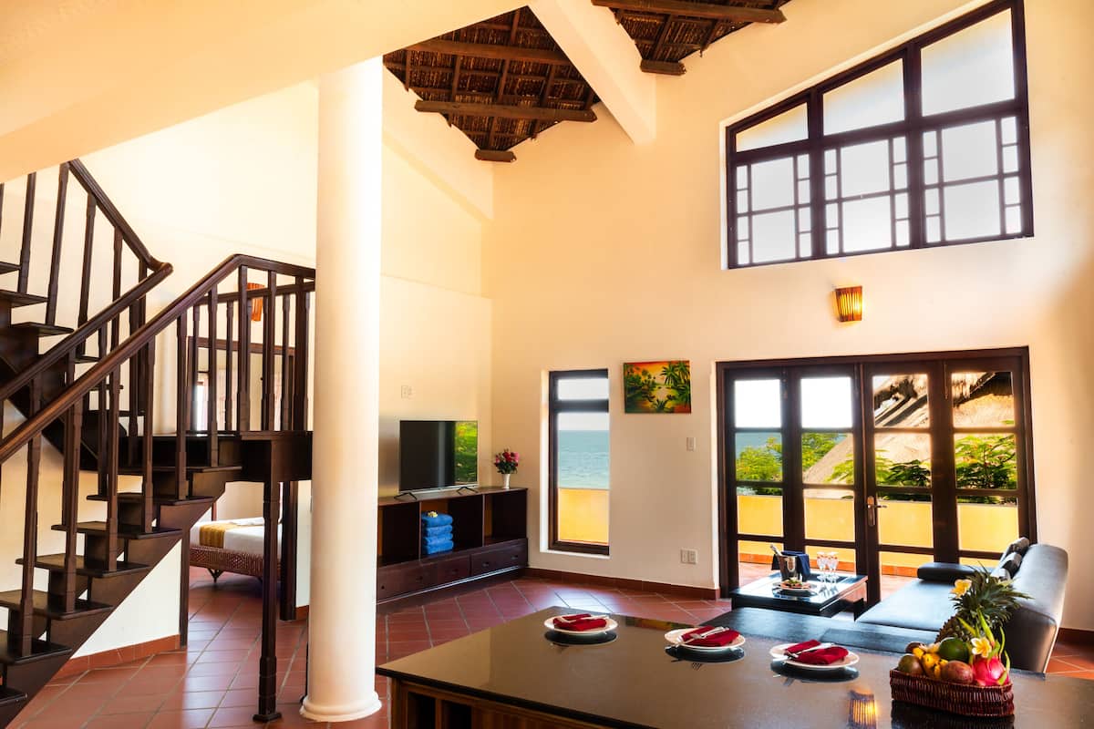 A spacious living area with a wooden staircase, a large window with a view of the ocean, and a dining table set with plates and a fruit basket.
