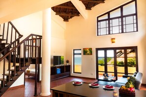 A spacious living area with a wooden staircase, a large window with a view of the ocean, and a dining table set with plates and a fruit basket. - White Sand Resort (Phan Thiet)