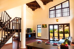 Amplia sala de estar con escalera de madera, un ventanal con vista al océano y una mesa de comedor dispuesta con platos y una cesta de frutas.
